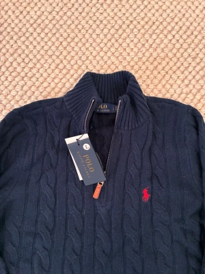 Mörkblå kabelstickad tröja Polo Ralph Lauren - Mörkblå kabelstickad tröja från Polo Ralph Lauren med halv dragkedja och hög krage. Tröjan har det klassiska röda broderade logotypen på bröstet och ribbstickade muddar. Perfekt för lager-på-lager och snygg till jeans.
