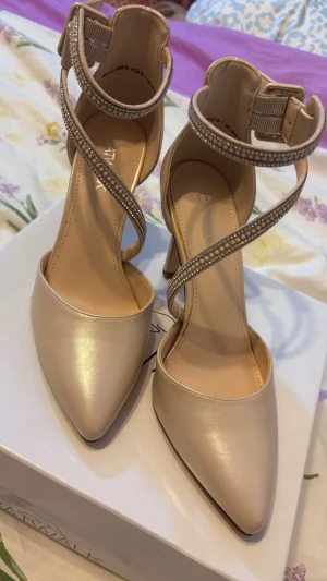 Beige pumps med strassdetaljer - Snygga pumps i beige från Catwalk med spetsig tå och höga klackar. Skorna har eleganta korsade remmar med strassdetaljer och justerbart spänne runt ankeln. Tillverkade i ett glansigt syntetmaterial som ger en lyxig känsla.