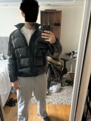 Svart pufferjacka från Moose Knuckles - Säljer en svart glansig pufferjacka från Moose Knuckles med hög krage och stora fickor framtill. Jackan har en patch med logga på ärmen och är tillverkad i ett blankt material som ger en modern look. Perfekt för kalla vinterdagar.