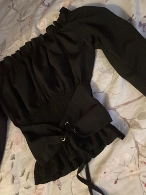Svart offshoulder topp med snörning - Trendig svart offshoulder topp med långa ärmar och snygg snörning i midjan. Toppen har en smockad överdel och volangkant nedtill som ger en feminin siluett. Perfekt för dig som vill ha en cool och stilren look.