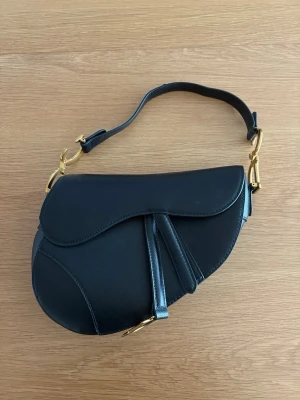 Svart Saddle Bag  - Väskan har en unik sadelform och kort axelrem, perfekt för att lyfta vilken outfit som helst. Klassisk och trendig design som sticker ut.