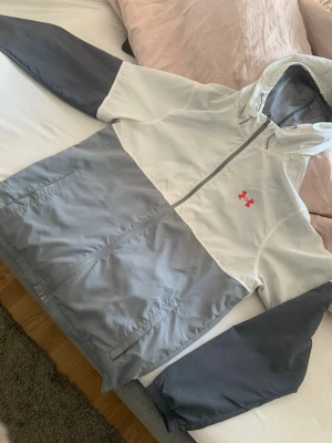 Gråvit vindjacka från Under Armour - Snygg vindjacka från Under Armour i grått och vitt med röd logga på bröstet. Jackan har huva, hel dragkedja och två sidofickor. Tillverkad i lätt och vindtätt material, perfekt för blåsiga dagar. Sportig design med meshfoder för extra komfort.