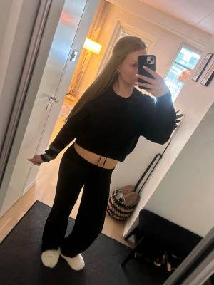 Svart croppad stickad tröja - Jag säljer en svart, kort tröja med avslappnad passform och långa ärmar. Tröjan har breda muddar vid ärmslut och nederkant, vilket ger den en snygg silhuett. Nedan finns en dragsko för att dra åt passformen vid höfterna. Perfekt för höst och vinter för en trendig och bekväm look.