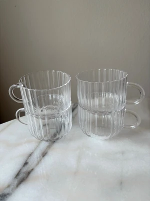 Räfflade genomskinliga glas-muggar - Fyra stilrena muggar i genomskinligt glas med räfflat mönster och rundade handtag. Perfekta för varma drycker och snygga att ha framme i köket. Muggarna är stapelbara och har en modern, minimalistisk design. 40cl koppar från Åhléns