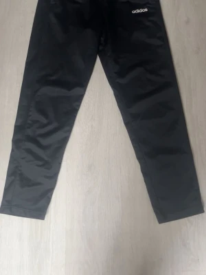 Svarta träningsbyxor från Adidas - Säljer ett par svarta träningsbyxor från Adidas med smal passform och diskret logga på ena låret. Byxorna är tillverkade i ett lätt material som passar perfekt för träning eller sportaktiviteter.
