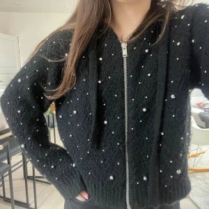 Svart kofta med pärlor och dragkedja - Säljer en svart, mysig kofta med dragkedja framtill och dekorativa pärlor över hela plagget. Koftan har huva, ribbade muddar och en lite lösare passform. Jättebra skick. Köptes för ca 600 från Zara.