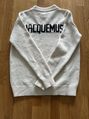 Vit stickad tröja från Jacquemus - Säljer en vit stickad tröja från Jacquemus med svart logotyp på ryggen. Tröjan har rund halsringning, ribbade muddar och nederkant. Perfekt för dig som gillar minimalistisk och trendig design.