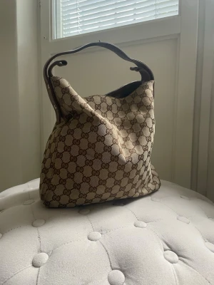 beige handväska - Säljer en klassisk beige handväska med det ikoniska GG-monogrammet över hela väskan. Köpt second hand, ej autentisk. Väskan har en mjuk, fyrkantig form och ett mörkbrunt handtag.