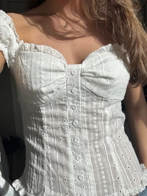 Vit broderad korsettopp - Supersöt vit korsettopp med broderade detaljer och knappar framtill. Toppen har offshoulder ärmar och spetskant längs urringningen. 
