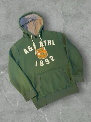 Grön hoodie Abercrombie & Fitch - Snygg grön hoodie från Abercrombie & Fitch med broderad text 'A&F ATHL 1892' och ett stort tygmärke på bröstet. Hoodien har en klassisk känguruficka, dragsko i huvan och kontrastfärgat innerfoder. Perfekt för en avslappnad och sportig stil.