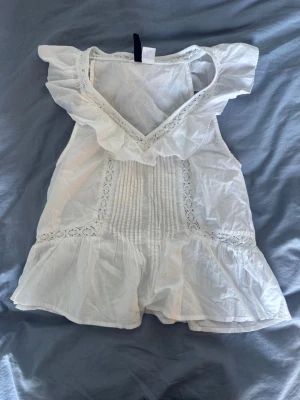 Vit volangblus med spetsdetaljer - Supersöt vit blus med volang vid v-ringningen och spetsdetaljer längs framsidan. Blusen är ärmlös och har en luftig passform med fina veck framtill. Perfekt för en fräsch och somrig look!