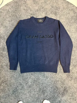 Blå stickad tröja Gran Sasso - Mörkblå stickad tröja från Gran Sasso med broderad logga och text på bröstet. Tröjan har rund halsringning, ribbstickade muddar vid ärmslut och nederkant. Perfekt för dig som gillar klassisk och stilren design.