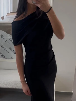 Svart oneshoulder långklänning - Elegant svart långklänning med oneshoulder-design och figurnära passform. Klänningen har en stilren och tidlös look som framhäver axlarna. Perfekt för dig som vill ha en klassisk och sofistikerad stil.