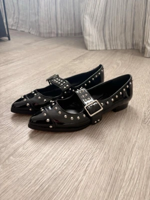 Svarta ballerinaskor med nitar - Säljer ett par svarta ballerinaskor i lackmaterial med silverfärgade nitar och en bred rem med spänne över foten. Skorna har spetsig tå och låg klack, vilket ger en edgy och trendig look. Perfekta för dig som vill sticka ut med detaljer.