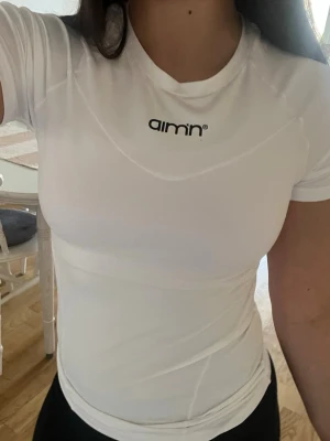 Vit träningströja från Aim'n - Säljer en vit, tight och kortärmad träningströja från Aim'n. Tröjan har rund halsringning och svart logga framtill. Perfekt för gymmet eller löpning, tillverkad i ett stretchigt funktionsmaterial.
