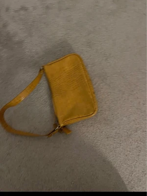 Gul axelväska i croco-mönster - Snygg gul axelväska med croco-mönstrad yta och enkel design. Väskan har en justerbar axelrem och är tillverkad i konstläder. Perfekt för dig som vill ha en färgklick till outfiten.