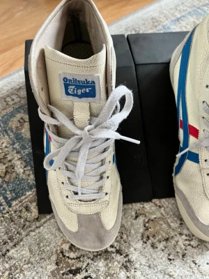 Onitsuka Tiger vita sneakers med blå ränder - Snygga vita sneakers från Onitsuka Tiger med klassiska blå och röda detaljer på sidorna. Skorna har en retro look med gräddvit ovandel i läder och mockadetaljer vid tån. Tjocka vita skosnören och Onitsuka Tiger-logga på plösen. Perfekta för dig som gillar vintagevibbar.