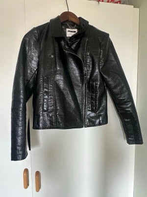 Svart croppad jacka i crocomönster - Snygg svart jacka med croppad passform och crocomönstrad yta. Jackan har snedställd dragkedja, klassisk krage och två fickor framtill. Perfekt för dig som vill ha en edgy look till din outfit.