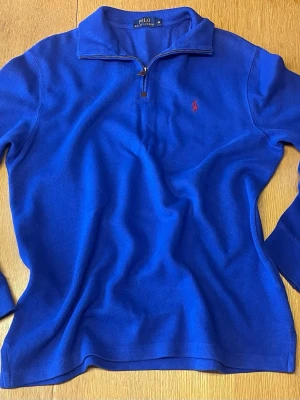 Blå halvzip tröja från Polo Ralph Lauren - Snygg blå långärmad tröja från Polo Ralph Lauren med halv dragkedja och klassisk röd logga broderad på bröstet. Tröjan har hög krage och är tillverkad i mjukt stickat material som passar perfekt till lager på lager. 70 cm längd 58 cm axel till axel.