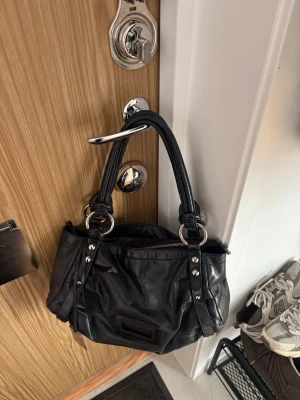 Svart axelväska från Coccinelle - Snygg svart handväska från COCCINELLE i mjukt konstläder med dubbla handtag och silverfärgade metalldetaljer. Väskan har en framficka och dekorativa remmar på sidorna. Perfekt storlek för att bära det viktigaste och passar till många olika stilar 🖤