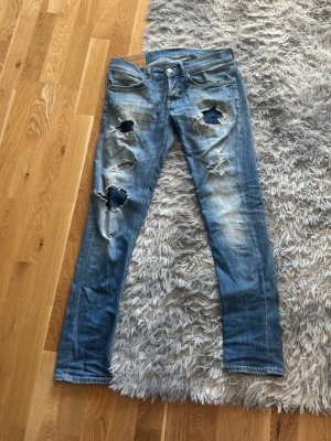 Blå slitna jeans från Dondup - Säljer ett par blå jeans från Dondup med flera stora slitningar och hål på framsidan. Jeansen har en smal passform och klassisk femficksdesign. Tillverkade i denim med snygg tvättad look och detaljerade sömmar.