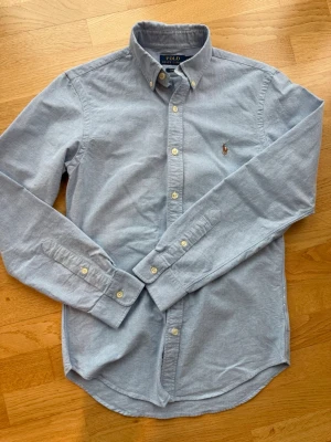 Ljusblå skjorta från Polo Ralph Lauren - Klassisk ljusblå skjorta från Polo Ralph Lauren med button-down krage och broderad logga på bröstet. Skjortan har långa ärmar, vita knappar och är tillverkad i mjuk bomull. Perfekt till jeans eller chinos för en clean look.