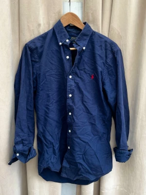 Marinblå skjorta från Polo Ralph Lauren - Klassisk marinblå skjorta från Polo Ralph Lauren. Skjortan har lång ärm och den ikoniska röda loggan på bröstet. Tillverkad i mjuk bomull för en bekväm känsla. Riktigt fin att ha när man är ute på middag eller ha under en väst till vardags🙌
