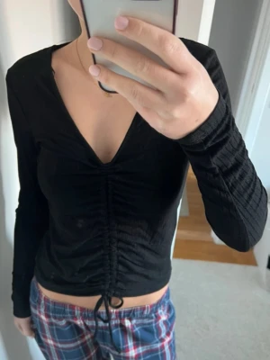 Svart ribbad topp med snörning - Trendig svart långärmad topp med ribbad struktur och snygg snörning framtill som ger en cool omlott-effekt. Djup v-ringning och croppad passform gör den perfekt till både jeans och kjol. Knappt använd. Märke: Lindex