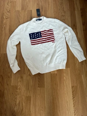Vit stickad tröja Polo Ralph Lauren - Vit stickad tröja från Polo Ralph Lauren med amerikansk flagga i rött, vitt och blått på bröstet samt broderade initialer 'RL'. Tröjan har rund halsringning och ribbade muddar vid ärmslut och nederkant. Perfekt för en klassisk och avslappnad stil.