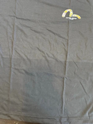 Svart Osaka Japan t-shirt - Svart t-shirt med ett litet tryck på bröstet där det står 'Osaka Japan' och ett färgglatt grafiskt motiv ovanför texten. Klassisk passform och rund halsringning. Perfekt till jeans eller shorts.