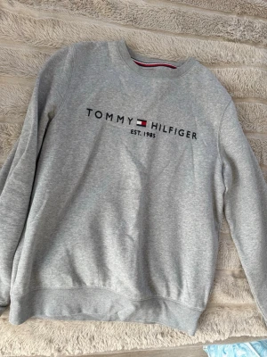 Grå sweatshirt Tommy Hilfiger - Snygg grå sweatshirt från Tommy Hilfiger med rund halsringning och broderad logga samt texten 'EST. 1985' på bröstet. Tröjan har ribbade muddar vid ärmslut och nederkant samt klassiska Hilfiger-detaljer i kragen. Perfekt för en avslappnad och stilren look.