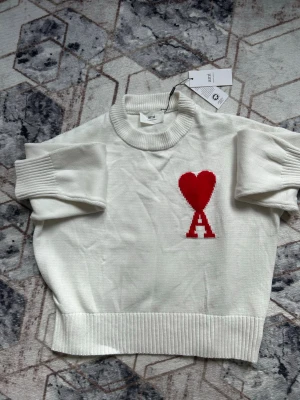 Vit stickad tröja från Ami Paris - Vit stickad tröja från Ami Paris med röd hjärt- och A-logga på bröstet. Tröjan har ribbade muddar vid ärmslut och nederkant samt en hög, ribbad rund halsringning. Oversized passform och mjukt material som känns lyxigt.