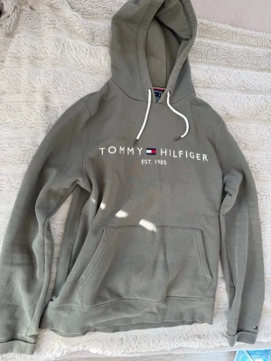 Olivgrön hoodie Tommy Hilfiger - Snygg olivgrön hoodie från Tommy Hilfiger med vit logga och text på bröstet samt klassiska flaggan. Hoodien har huva med vita snören och en stor magficka framtill. Tillverkad i mjuk bomullsblandning, perfekt för en avslappnad och trendig look.