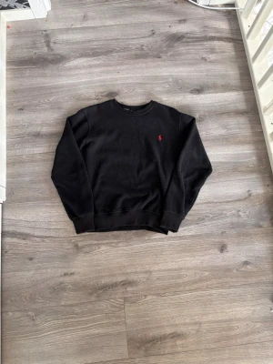 Svart sweatshirt från Ralph Lauren - Klassisk svart sweatshirt från Ralph Lauren med rund halsringning och den ikoniska röda logotypen broderad på bröstet. Tröjan har ribbade muddar vid ärmslut och nederkant. Perfekt för en avslappnad och stilren look.