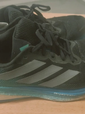 Svarta Adidas Dreamstrike 2 sneakers - Säljer ett par svarta Adidas Dreamstrike sneakers med klassiska tre ränder i silver på sidan. Skorna har mesh-ovandel för bra ventilation och blågrön sula som ger en sportig vibe. Perfekta för dig som gillar stilrena och bekväma sneakers. Nypris 1799