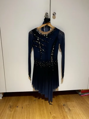 Mörkblå konståknings klänning  - Snygg mörkblå tävlings klänning med långa meshärmar och transparent mesh upptill. Klänningen har massor av glittriga stenar och pärlor i silver och guld, samt asymmetrisk. Perfeckt för program med intensiv/ mäktig låt