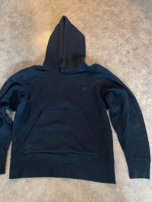 Svart hoodie från Acne Studios - Säljer en svart hoodie från Acne Studios med det klassiska smiley-märket på bröstet. Hoodien har stor huva, känguruficka och ribbade muddar. Tillverkad i mjuk bomull för skön känsla och stilren look.