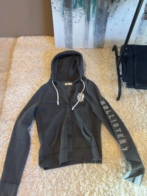 Mörkgrå hoodie från Hollister - Snygg mörkgrå hoodie från Hollister med knappar framtill, vita dragsnören i huvan och tryck på ena ärmen samt bröstet. Tillverkad i mjukt bomullsmaterial och har två fickor framtill. Perfekt för en avslappnad och trendig look.