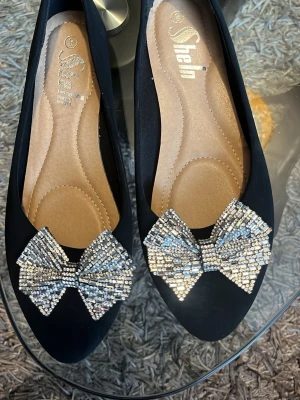 Svarta ballerinaskor med strassrosett - Säljer ett par svarta ballerinaskor från Shein med stora glittriga rosetter framtill, täckta av silvriga strasstenar. Skorna har rundad tå och mjuk innersula i beige. Perfekta för dig som vill ha något extra till outfiten.