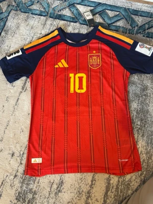 Spaniens matchtröja Lamine Yamal #10 - Röd och marinblå matchtröja från Adidas för Spaniens landslag med gula detaljer, nummer 10 och Lamine Yamal på ryggen. Player version jätte bra kvalité med patch på vänster och höger axel 