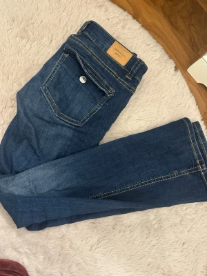 Blå jeans från Perfect Jeans - Säljer ett par klassiska blå jeans från Perfect Jeans med snygga kontrastsömmar och en unik ficka med lock bak. Jeansen har normal midja och raka ben. Perfekta till vardagslooken och tillverkade i mjukt denimtyg.