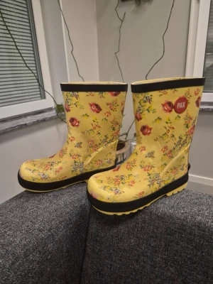 Gula blommiga stövlar från PAX 28 - Snygga gula stövlar från PAX med färgglatt blommönster och svarta detaljer runt sula och kant. Tillverkade i gummi och perfekta för regniga dagar. Stövlarna har en klassisk hög modell och sticker ut med sin lekfulla design.