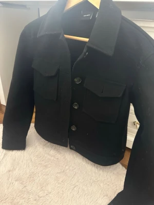 Svart kappa från Vero Moda - Stilren svart kappafrån Vero Moda med klassisk krage och fyra stora fickor framtill. Jackan har raka ärmar och stängs med knappar. Materialet ser ut att vara tjockt och mjukt, perfekt för lager på lager. Enkel och trendig design som passar till det mesta.