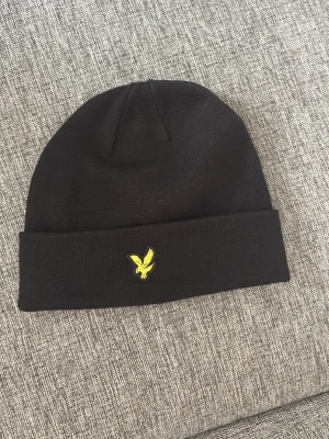 Svart mössa från Lyle & Scott - Säljer en svart ribbstickad mössa från Lyle & Scott med klassisk gul logga framtill. Mössan har uppvikt kant och är tillverkad i mjukt material som håller dig varm. Enkel och stilren design som passar till det mesta.