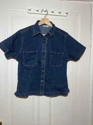 Kortärmad jeansskjorta i denim - Snygg mörkblå jeansskjorta i denim med korta ärmar och två bröstfickor. Klassisk krage och knappar framtill. Perfekt för dig som gillar en avslappnad och trendig stil.