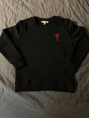 Svart sweatshirt från Ami Paris - Stilren svart sweatshirt från Ami Paris med det ikoniska röda hjärtat och bokstaven A broderat på bröstet. Tröjan har rund halsringning, ribbade muddar och är tillverkad i mjukt material som passar perfekt till en avslappnad look.