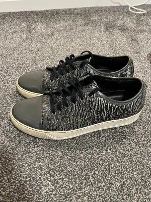Lanvin Sneakers – Svart/Vit, Storlek 42-43 - Snygga svarta sneakers från Lanvin med stickad ovandel och vita detaljer på sulan. Tåpartiet är i blankare material och skorna har svarta skosnören. Zebra-liknande mönster gör dem riktigt unika.  Perfekta för dig som gillar en stilren och modern look – passar lika bra till jeans som streetwear.  Skick: Bra, lite små heeldrag och ett litet märke (knappt synligt på bild). Pris 1899kr! Storlek: 42-43