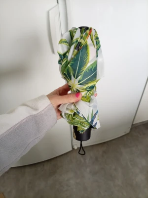 Paraply med tropiskt mönster - Säljer ett hopfällbart paraply med vitt tyg och färgglatt tropiskt mönster av gröna blad och blå papegojor. Paraplyet har svart handtag med handledsrem och är perfekt att ha i väskan när regnet överraskar. Materialet är syntetiskt och lätt att torka av.