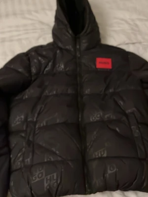 Svart pufferjacka från Hugo - Snygg svart pufferjacka från Hugo med diskret logomönster över hela jackan och röd patch med Hugo-logga på bröstet. Jackan har huva, dragkedja framtill och röda detaljer på insidan. Perfekt för kalla vinterdagar och riktigt trendig look.