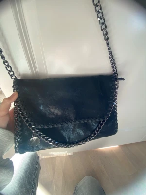 Svart axelväska med kedjedetaljer - Snygg svart axelväska med grova silverfärgade kedjor längs kanterna och som axelrem. Väskan har lock med magnetknäppning och innerfack med dragkedja. Materialet ser ut som glansig konstläder och väskan har en rund metallbricka som detalj.